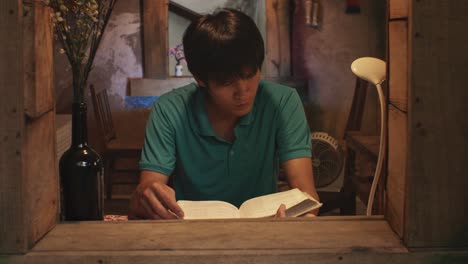 looking through an open window at a young asian male reads a book at a desk in a rustic room