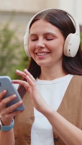 mujer joven encantada con auriculares inalámbricos eligiendo escuchar música sosteniendo un teléfono inteligente en la calle