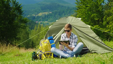 Un-Joven-Turista-Usa-Una-Computadora-Portátil-Mientras-Acampa-Cerca-De-Una-Carpa.