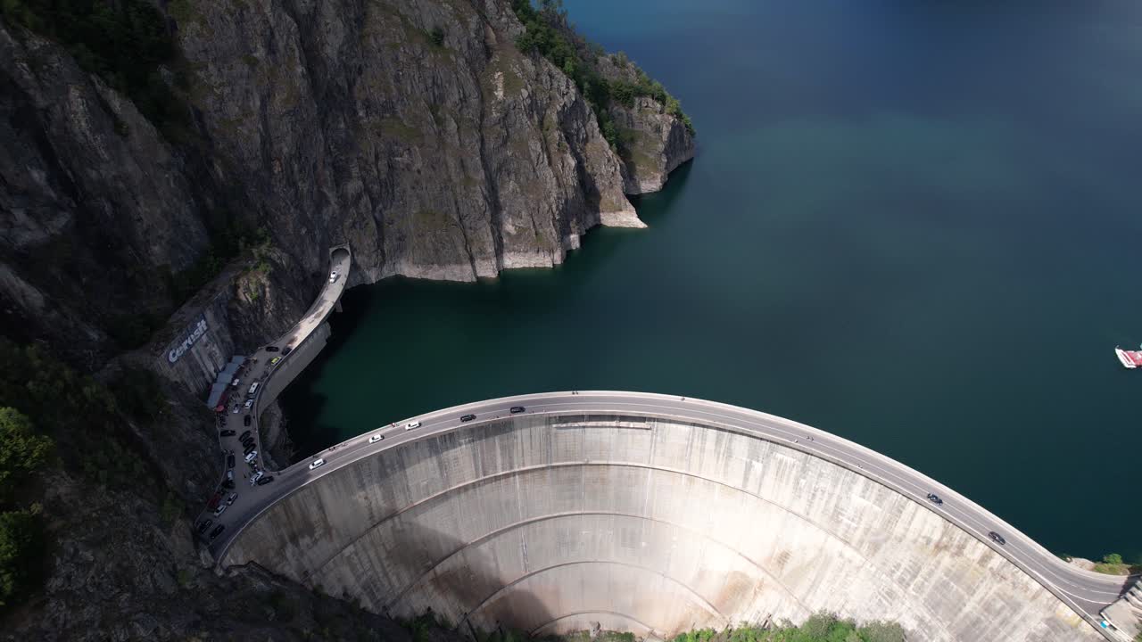 Rising Aerial shot of Vidraru Dam in Romania, captured at an angle while circling left. Blue lake, dramatic curves, and green mountain backdrop in 4K.