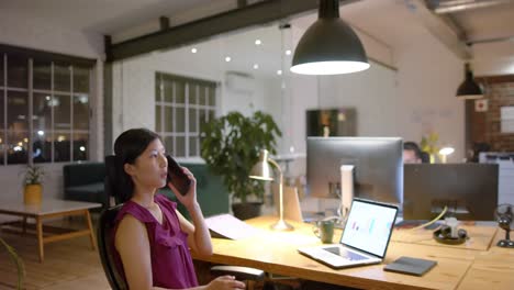 Happy-asian-casual-businesswoman-using-laptop-talking-on-smartphone-in-office-at-night,-slow-motion