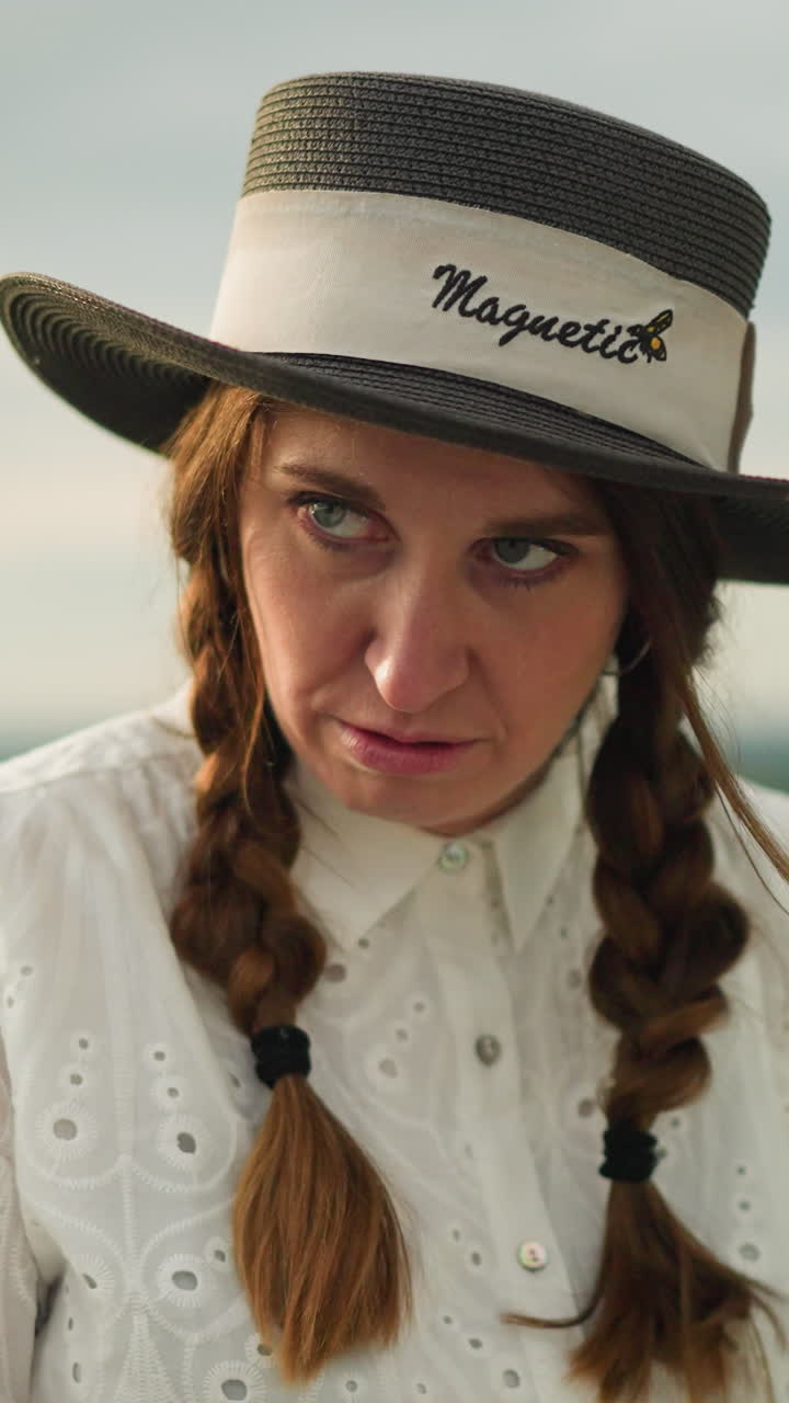 클로즈업은 색 드레스를 입고, 머리카락을 고, 넓은 가장자리 모자를 입은 여자의 강렬한 표정을 포착합니다.