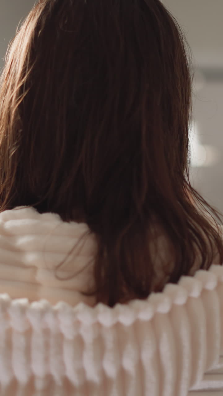 넓은 욕실에 서있는 여성. 미용 치료 후 후드와 함께 욕실 로브에 서있는 갈색 여성은 욕실 뒷면 램프와 함께 거울 근처에 서 있습니다.