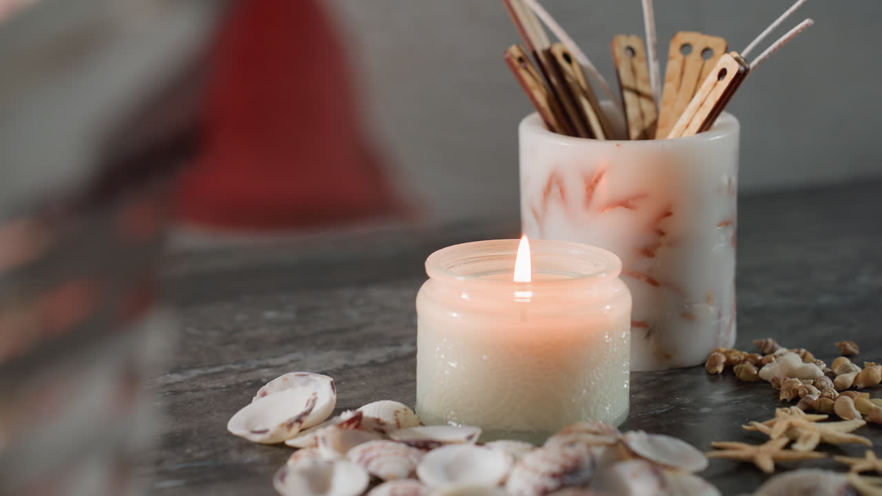 Close up of white candle burning brightly in glass jar with seashells and starfish on dark marble table next to woven basket with red cloth