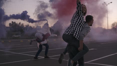 Dos-Parejas-Jóvenes-Locas-Con-Bombas-De-Humo-De-Colores-En-Las-Manos-En-La-Zona-De-Estacionamiento-Vacía-Afuera