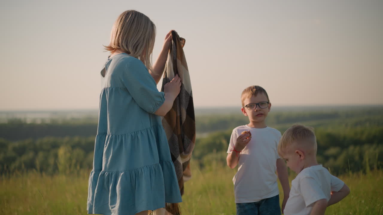 A mother in a blue dress is seen carefully folding a scarf in a grassy field, preparing to hand it to her younger son who stands close by. His older brother, also in a white top, watches attentively