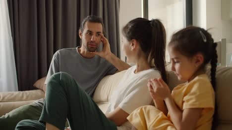 Close-up-shot-of-a-happy-father-with-long-hair-and-stubble,-a-brunette-guy-communicates-with-his-girlfriend,-the-mother-of-their-daughter,-on-a-brown-sofa-sofa-in-a-modern-room