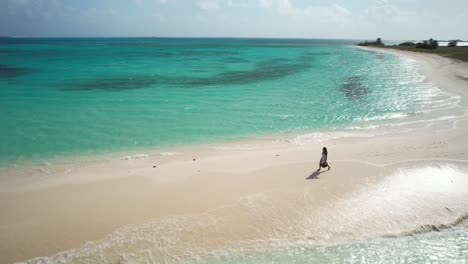 Drohnenaufnahme-Einer-Frau,-Die-Bei-Hintergrundbeleuchtung-Den-Sonnenuntergang-Am-Weißen-Sandstrand-Spaziert,-Meerwasserspritzer-Und-Absturz-Auf-Der-Sandbank