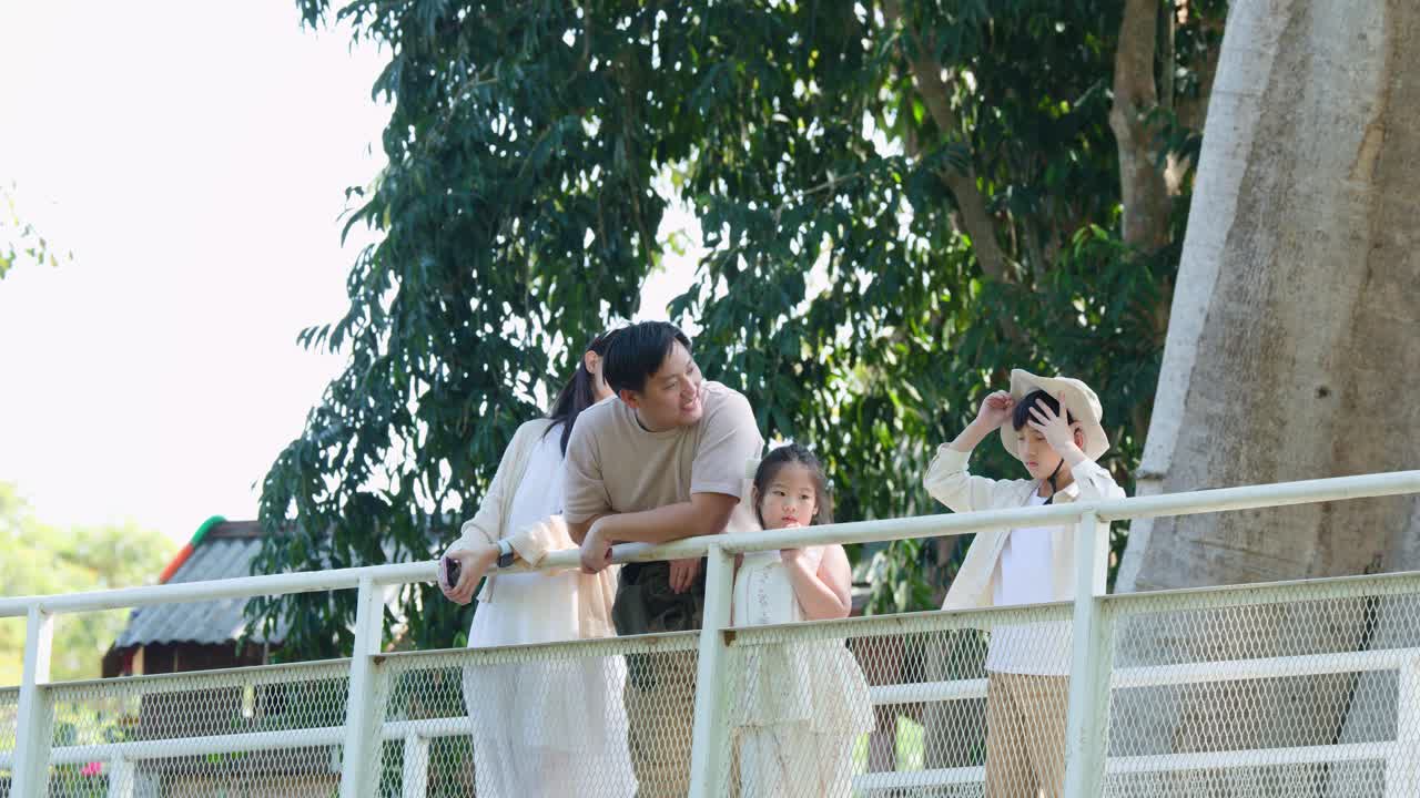 ครอบครัวสุขสันต์สี่คนยืนอยู่บนสะพาน ยิ้มแย้มและสร้างความสัมพันธ์ภายใต้แสงอาทิตย์ธรรมชาติ