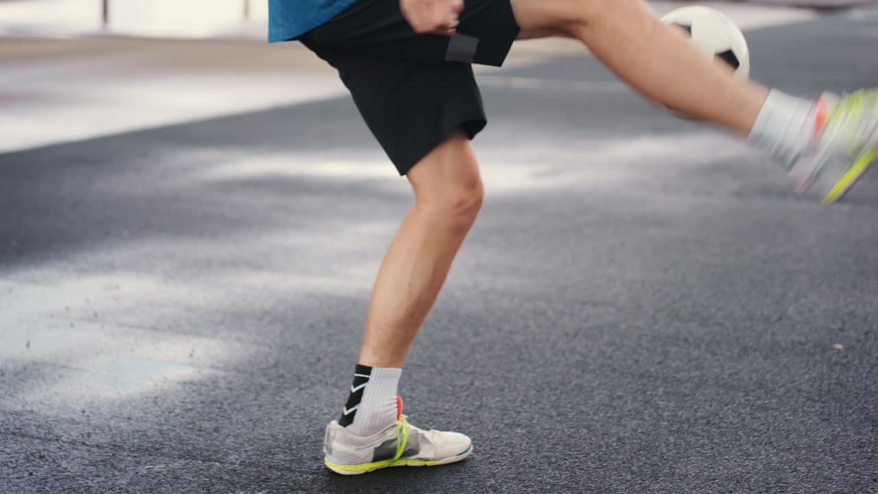 Männerbeine jonglieren Fußball auf regnerischem Asphalt, Nahaufnahme einer Trainingseinheit mit Neon-Sneakers und Knöchelsocken, Pfützen und reflektierende Oberfläche, kontrollierte Berührungen und leichte Spritzer, rhythmische Beinarbeit