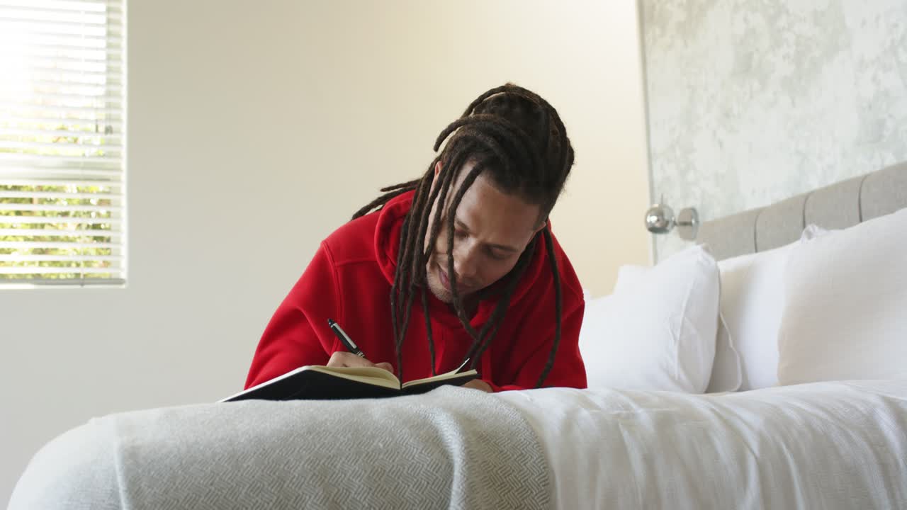 Uomo che scrive con una penna su un quaderno, seduto sul letto in camera da letto, mentre guarda fuori dalla finestra e riflette.