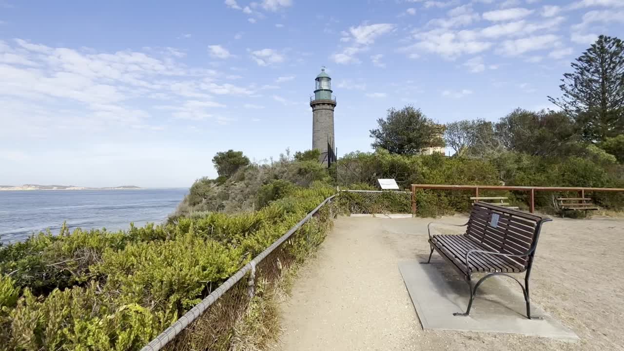 Static shot showing a bench near the sea with Queenscliff Black Lighthouse in the background on the Bellarine Peninsula, Australia, under a clear sky