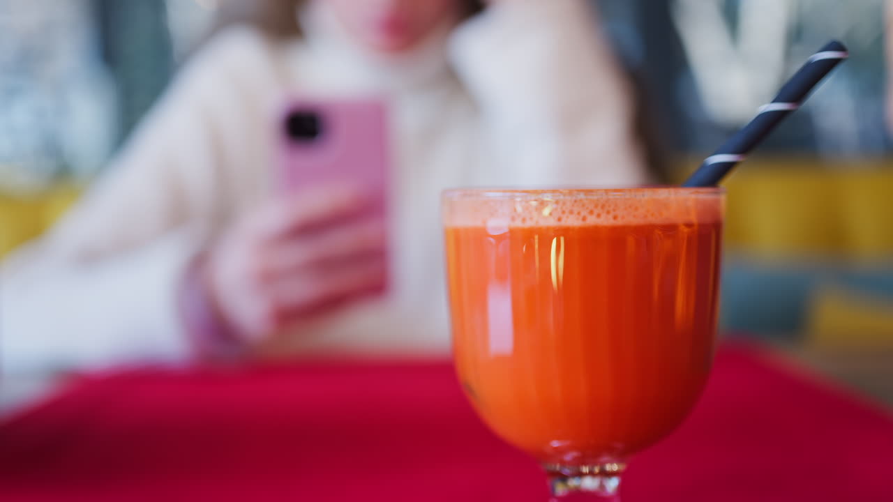 Close up of an orange and carrot juice in a glass with a black straw on a red table cloth with a woman on the background
