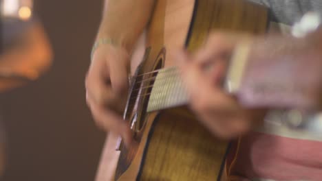 primer plano de un músico profesional tocando acordes en una guitarra occidental con un baterista borroso en el fondo durante una sesión en vivo con cálidas luces de estudio