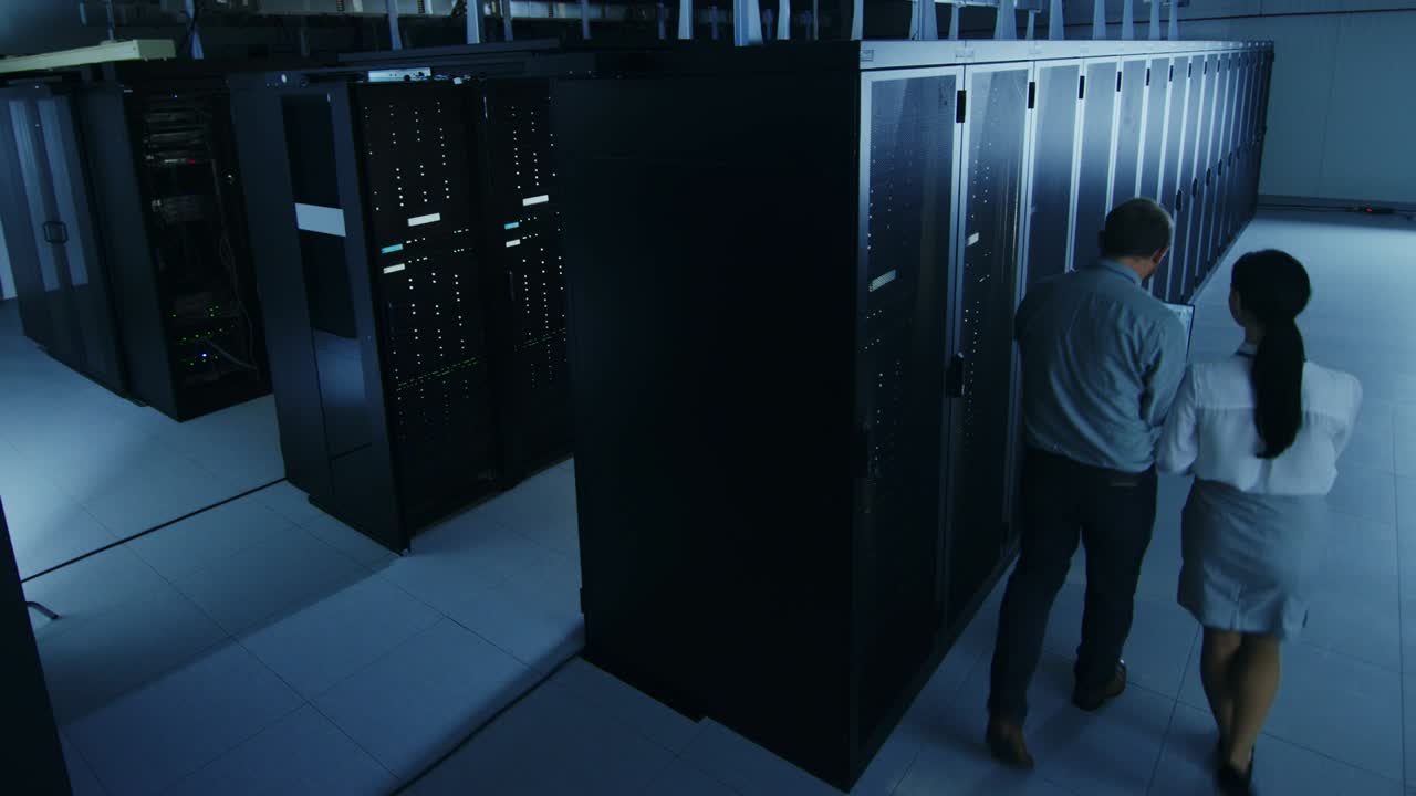 por encima de una toma amplia de un administrador de ti barbudo con gafas con una computadora portátil y un joven colega técnico hablando en el centro de datos mientras camina junto a los estantes de servidores. ejecutando diagnósticos o mantenimiento.