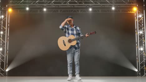 cuerpo lleno de adolescente asiático con guitarra que tiene dolor de cabeza mientras está de pie en el escenario