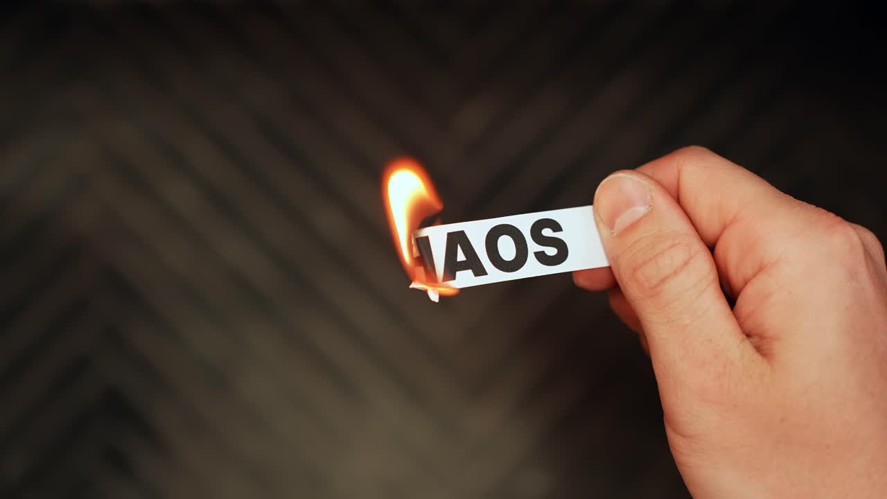 Primo piano di una mano che tiene un'etichetta di carta con la scritta “CHAOS” mentre il bordo prende fuoco, una metafora drammatica per lo stress e il disordine con spazio per il testo.