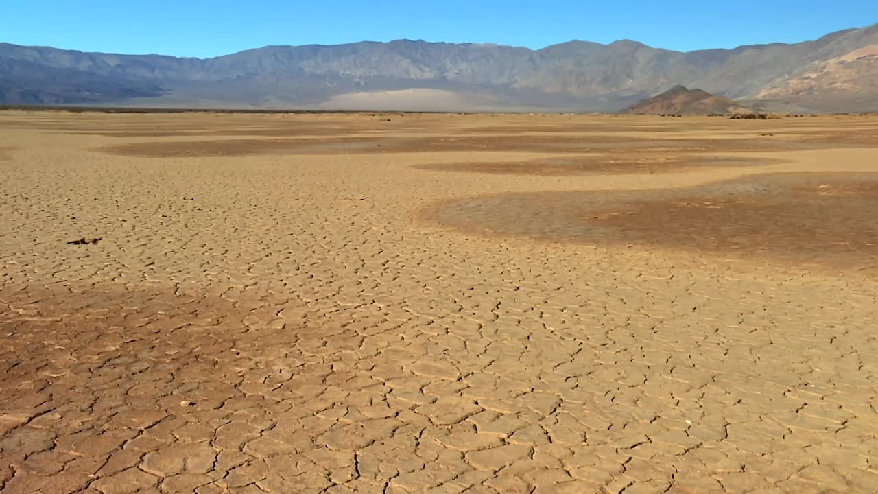 Expansive cracked mud surface with polygonal patterns stretches across desert plain under clear sky, with distant mountains in Death Valley National Park, California, USA