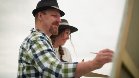 a happy artist holding a paint palette and working on a large canvas in an outdoor .he smiles as he paints, while beside him, a woman in a white dress and hat stands, joyfully observing his work