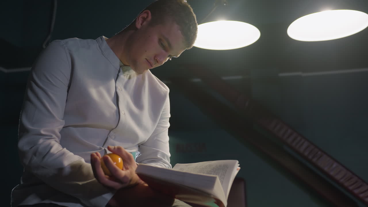 toma de primer plano de un caballero con camisa blanca sentado en una mesa de billar verde, leyendo un libro mientras tira una pelota de billar en la mano. expresión enfocada, iluminación tenue y fondo de mal humor