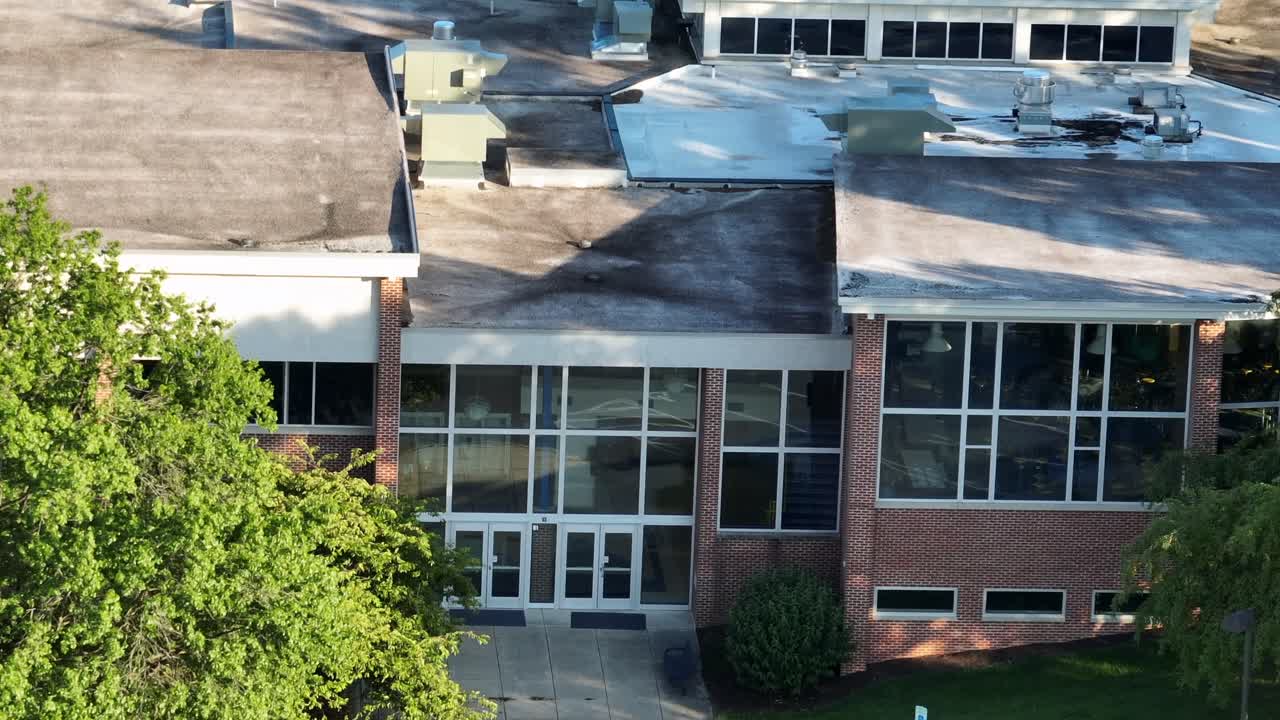 American school with red brick and large windows at golden sunset in summer holidays. Aerial zoom shot. Pipelines and air conditioner on roof. Closed public school in Virginia, USA