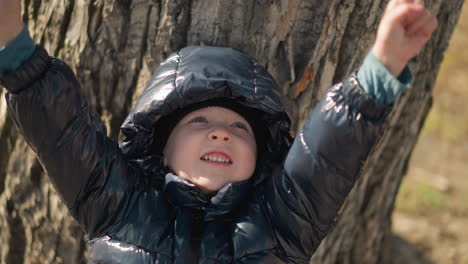 un niño apoyado en un gran tronco de árbol, mirando hacia arriba con la boca abierta de emoción, sus brazos levantados, y está vestido con una chaqueta negra brillante