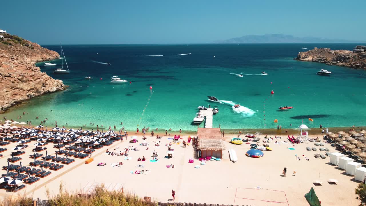 Aerial drone shot revealing Super Paradise Beach in Mykonos, Greece during a clean sky sunny day with tourists and water sports
