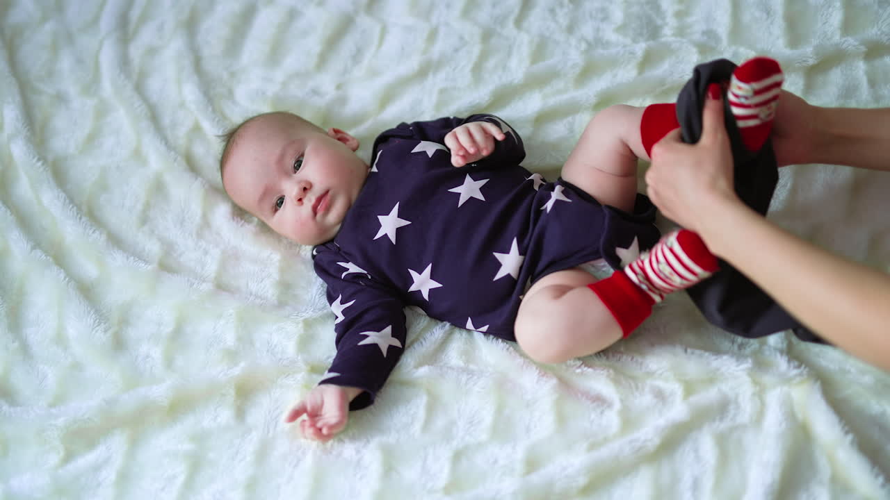 Loving mother puts black pants on little baby boy. View from above. White plaid backdrop.