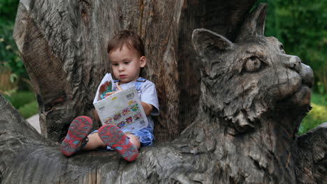 serious baby boy sitting on top of a wooden cat sculpture. adorable kid turns the pages of a children book.
