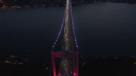 Hermoso-Puente-Rojo-Sobre-El-Bósforo-En-Luz-Roja-Por-La-Noche-Con-Tráfico-De-Camiones,-Vista-Aérea-De-Pájaros-Inclinada-Hacia-Abajo