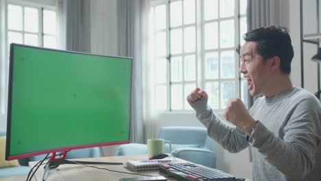 asian man in long sleeved t-shirt with green screen desktop computer being happy successful in work while working at home.