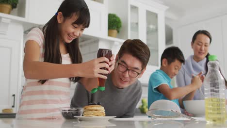 happy asian parents in kitchen cooking with son and daughter, daughter putting syrup on pancakes