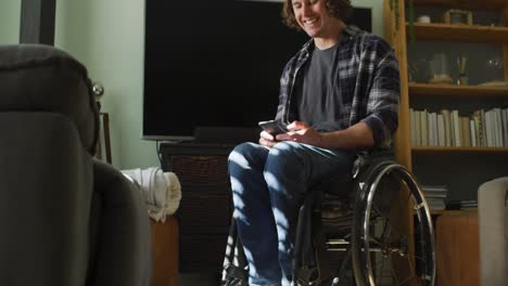 hombre discapacitado caucásico feliz en silla de ruedas usando un teléfono inteligente en la sala de estar