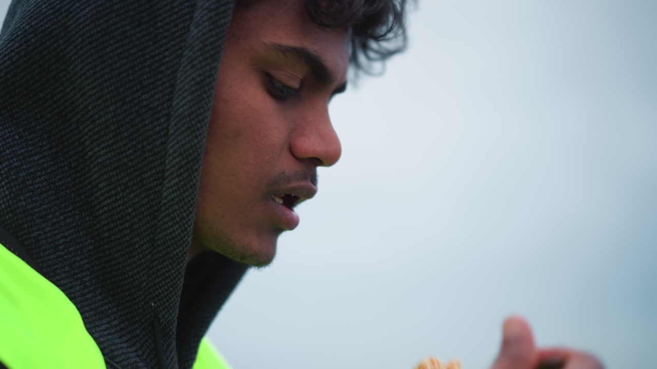 Young male worker in dark hoodie and neon reflective vest looking down thoughtfully in cold outdoor setting, showing focus, during work in urban environment under overcast sky