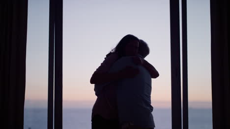 Silueta-Feliz-Pareja-De-Ancianos-Abrazándose-En-La-Habitación-Del-Hotel-Mirando-Por-La-Ventana-Al-Atardecer-Disfrutando-De-Un-Exitoso-Estilo-De-Vida-De-Jubilación-En-Vacaciones-Compartiendo-Conexión-Romántica