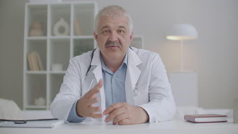 portrait of male therapist is speaking to camera sitting at table in his office in clinic giving recommendations