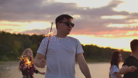 el hombre brutal está saltando en baile con grandes luces bengalíes en la playa de arena con sus amigos. esta es una maravillosa noche de verano en la fiesta al aire libre.