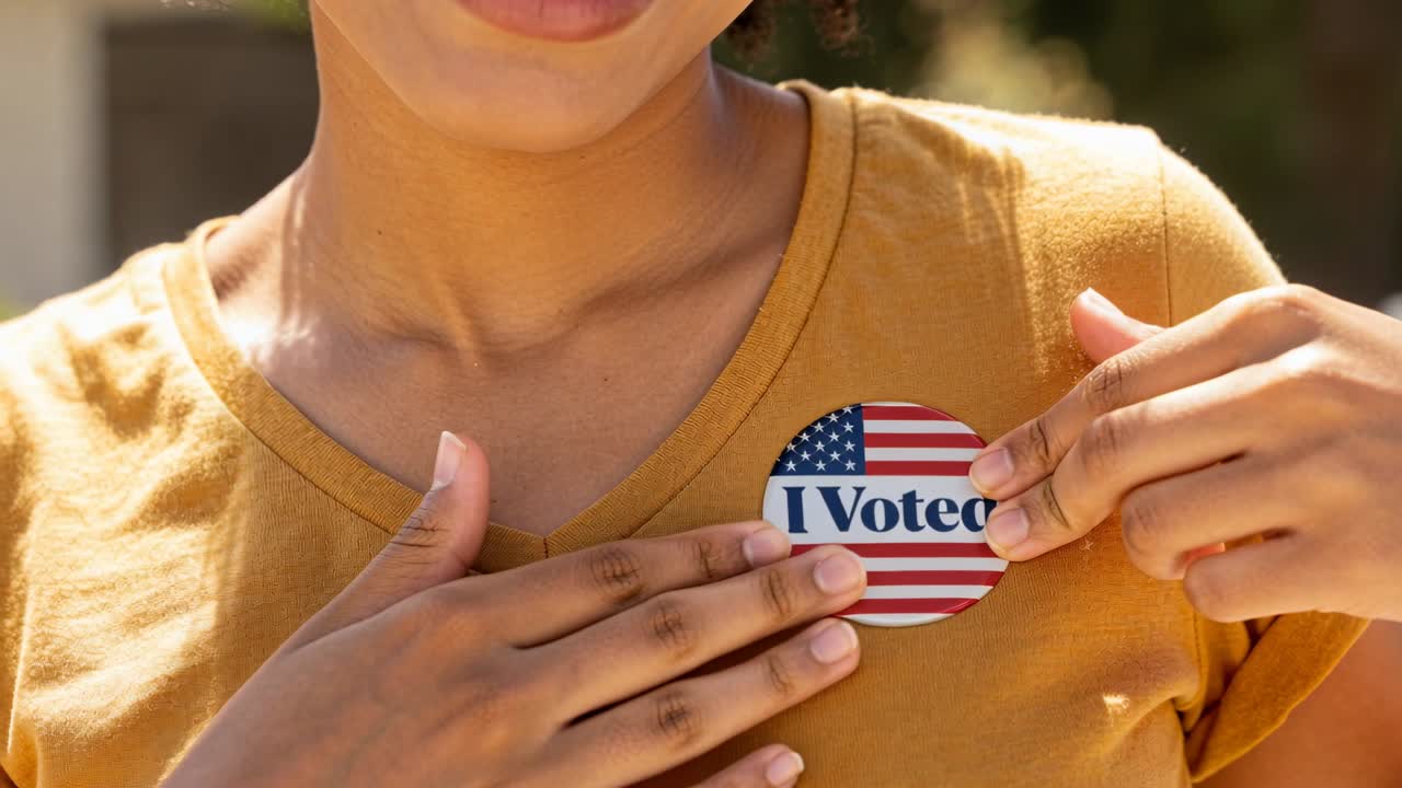 Placing woman aligning pressing I Voted sticker on mustard V-neck shirt on front porch after voting