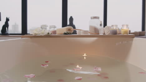 camera focuses on a white bathtub with water, flowers and candles and wooden table with bathroom elements