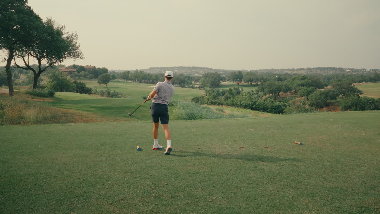 Erkek bir golfçü, tee kutusundan 3-wood ile ağır çekimde vuruş yapıyor, ardından sağlam bir vuruşun ardından sakince eğilip tee'yi alıyor. Güneşli bir sahada özgüveni, ritmi ve vuruş sonrası rutinini yansıtıyor.