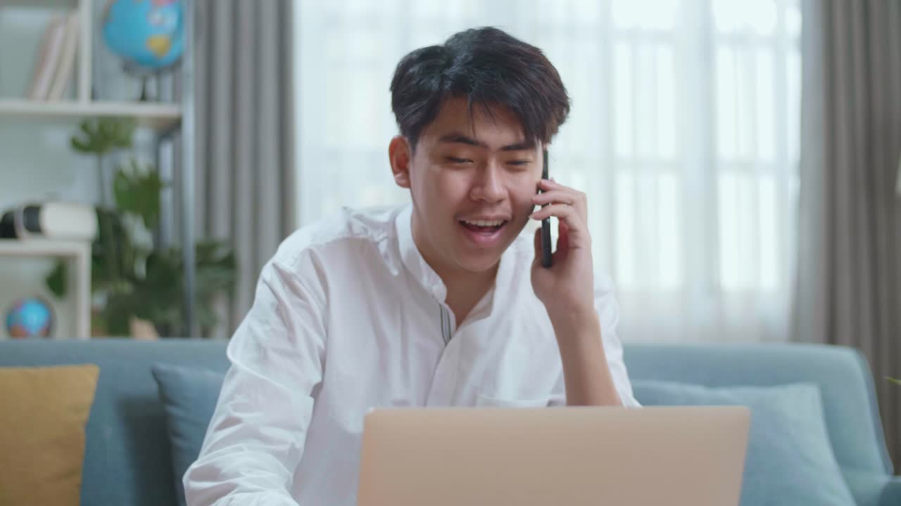 Smiling Asian Man Answering The Call On Smartphone While Using Laptop Computer In The Living Room
