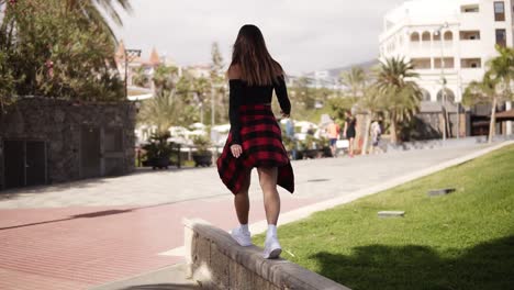 stylish girl is stepping by parapet, balancing. fashionable tourist in casual clothes walking along the tropical trees park, resort area on parapet. elegant stylish woman enjoy the time. rare view