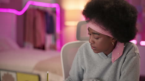 a young woman with an afro and a pink headband is working on her laptop. she is sitting at a desk in an office and has a pencil in her hand. she looks focused and determined.