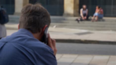 Defokussierte-Aufnahme-Eines-Mannes,-Der-Telefoniert,-Während-Er-Auf-Der-Straße-In-Oxford-01ox-Sitzt