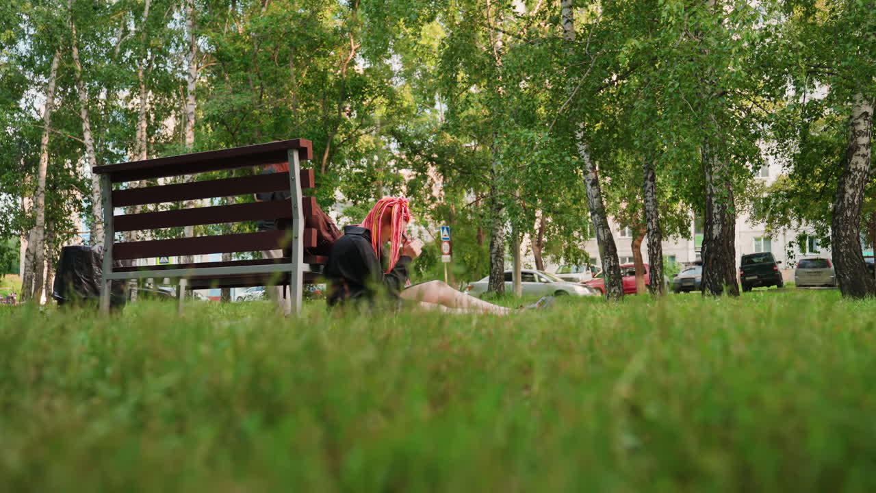 햇빛이 비치는 공원의 벤치 옆에 누워 있는 백인 여성, 분홍색과 노란색 땋은 머리가 어깨에 드리워져 있고, 넓은 잔디밭이 전경을 이루며 자작나무가 배경을 이루고, 부드러운 여름 햇살과 함께 편안하면서도 외로운 분위기