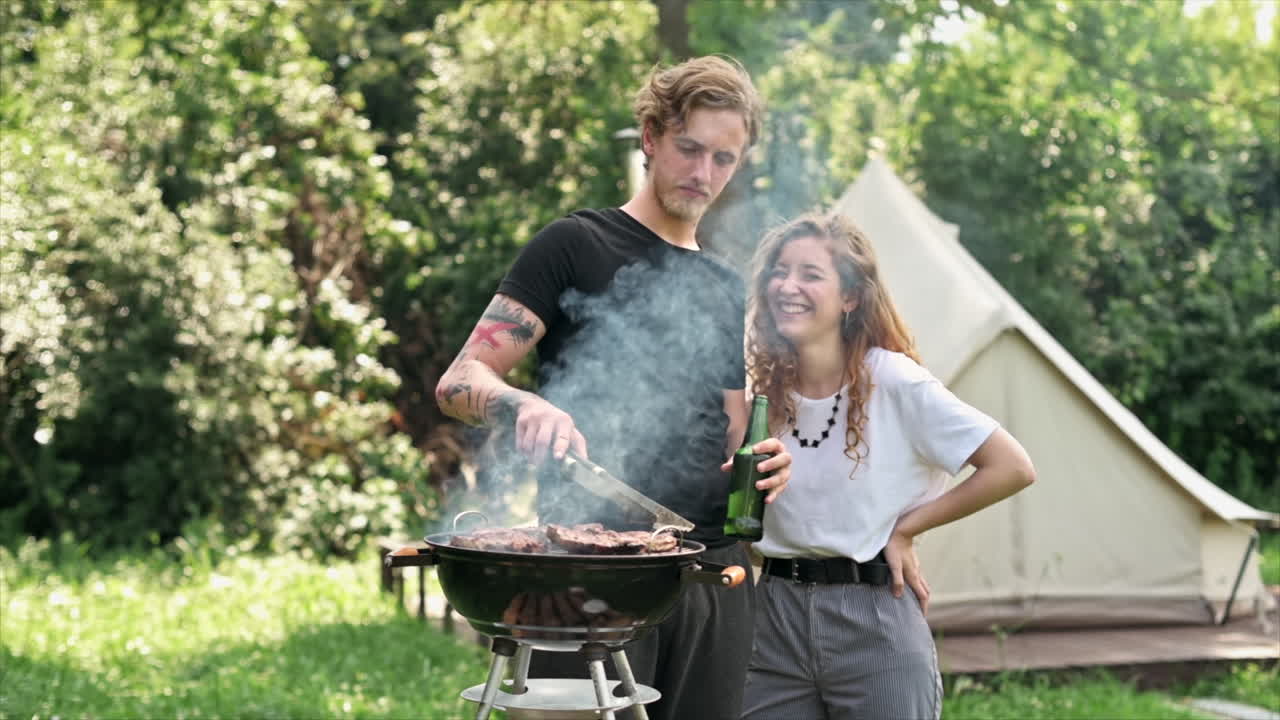 Giovane coppia che frigge carne alla griglia e beve birra, ridendo. Tanto verde intorno. Glamping. Slow motion