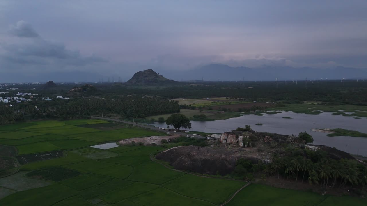 This atmospheric aerial footage captures a large wind farm set in a dynamic landscape. In the foreground, a rocky outcrop overlooks a large lake or reservoir with patches of vegetation