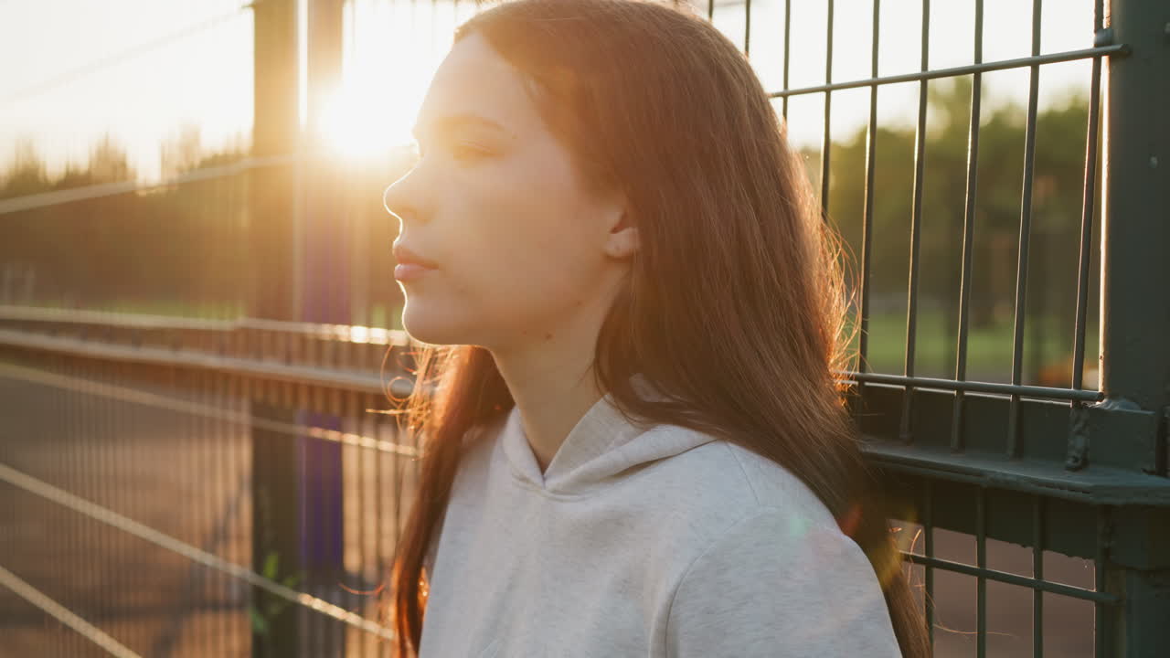 한 여인이 일몰의 빛에 스포츠 경기장의 울타리 에 서 있습니다. 젊은 여성은 훈련 후 휴식을 취하는 동안 문제에 대한 생각에 몰두합니다. 공허함의 느낌.