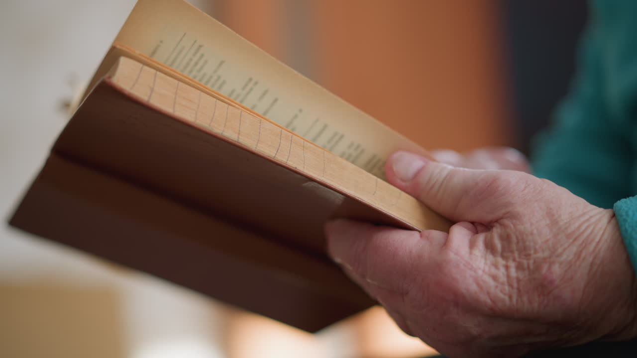 Close-up of lonely pensioner holding brown book in hands, reading in peaceful setting. Elderly person in green teal sweater, quiet moment of concentration, with light softly illuminating the scene