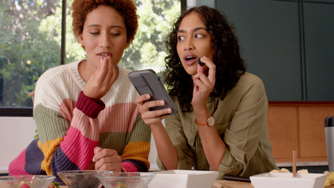 female friends laughing and chatting while looking at smartphone in kitchen